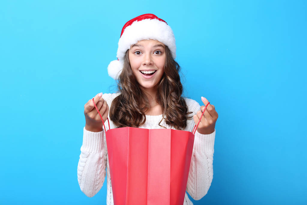 Beautiful girl in santa hat holding red shopping bag on blue background - Photo, Image