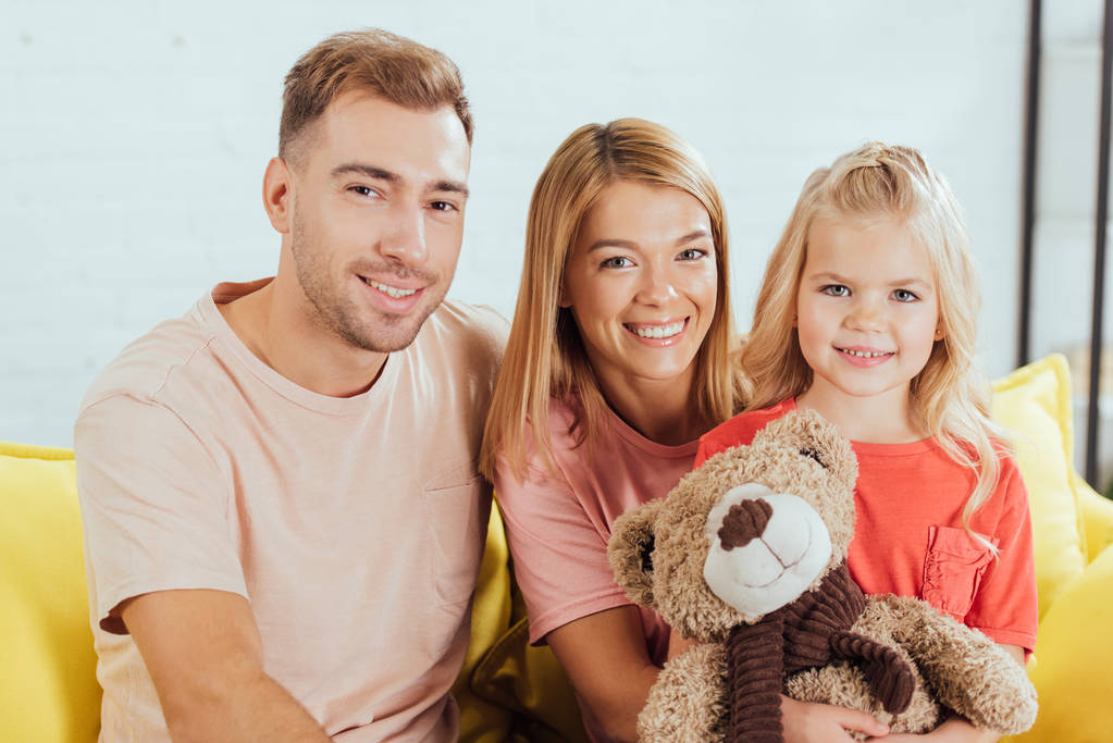 Lächelnde Eltern, die auf der Couch sitzen, in die Kamera schauen und ihre Tochter einen Teddybär in der Hand halten - Foto, Bild
