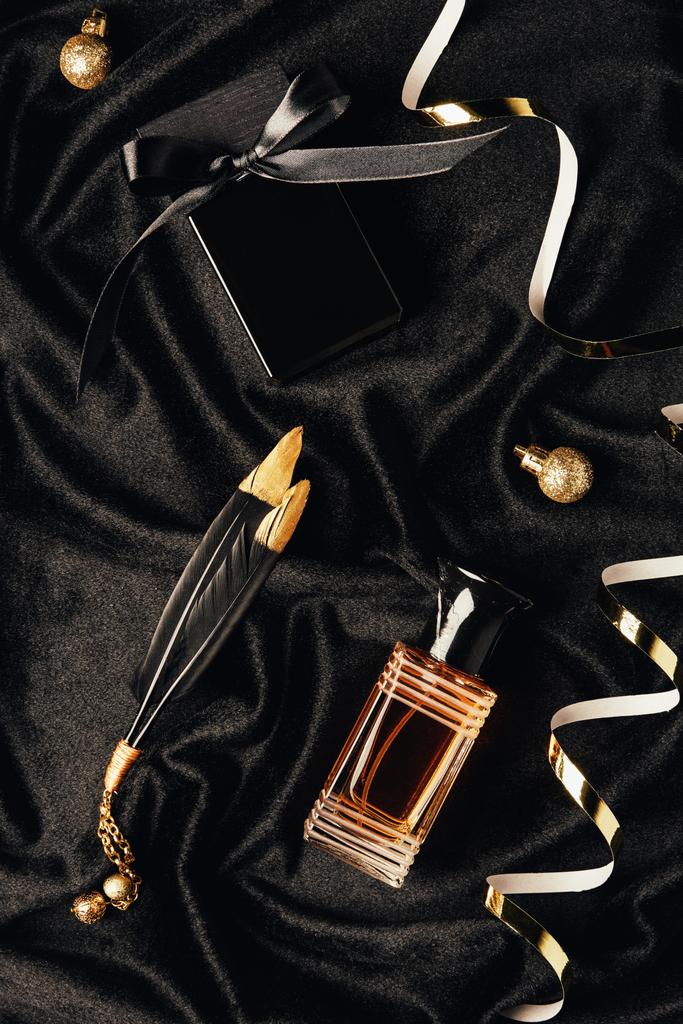 bovenaanzicht van parfums, decoratieve veren, gouden linten en kerstballen op zwarte stof - Foto, afbeelding