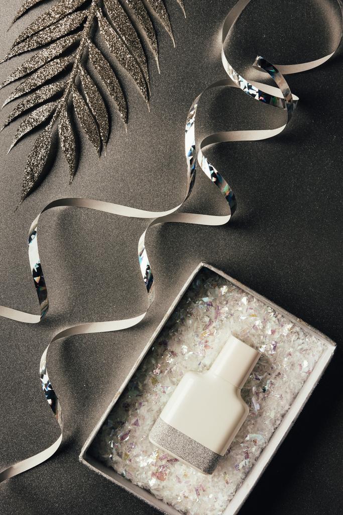 парфюмерная бутылка в коробке, серебряные ленты и блестящие декоративные листья на сером
  - Фото, изображение