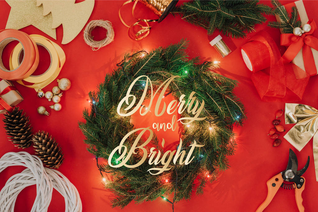 çam ağacı dalları, Noel ışıkları ve şeritleri için el yapımı Noel çelenk ile "neşeli ve parlak" harflerle kırmızı zemin üzerinde düz lay  - Fotoğraf, Görsel