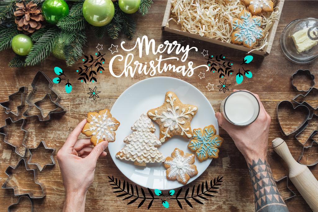 cropped shot of man holding homemade christmas cookie and glass of milk on wooden surface with "merry christmas" lettering - Photo, Image