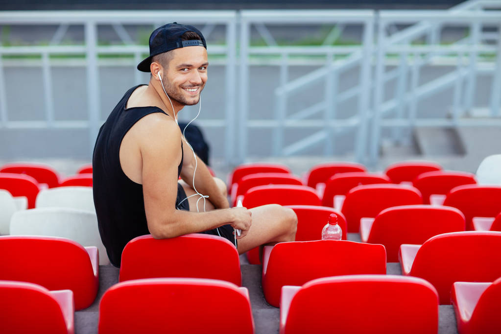 Jovem atleta sorrindo sentado em assentos de estádio e ouvindo música. relaxe conceito
 - Foto, Imagem