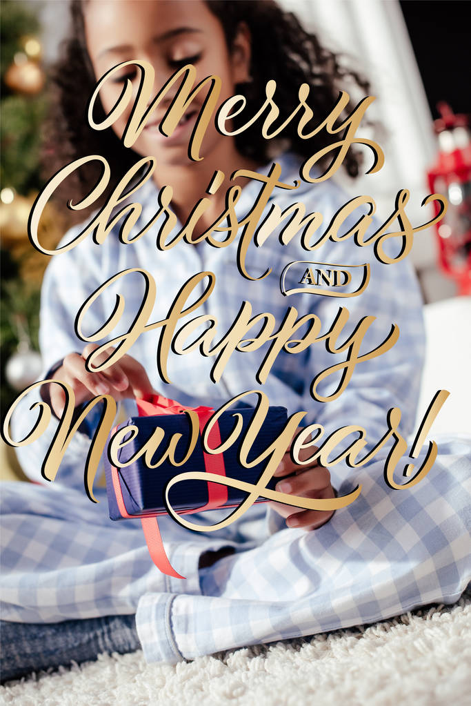 επιλεκτική εστίαση του χαμογελώντας αξιολάτρευτο παιδί αφροαμερικάνων στις πυτζάμες ανοίγοντας το δώρο των Χριστουγέννων στο σπίτι με «καλά Χριστούγεννα και Ευτυχισμένο το νέο έτος» γράμματα - Φωτογραφία, εικόνα