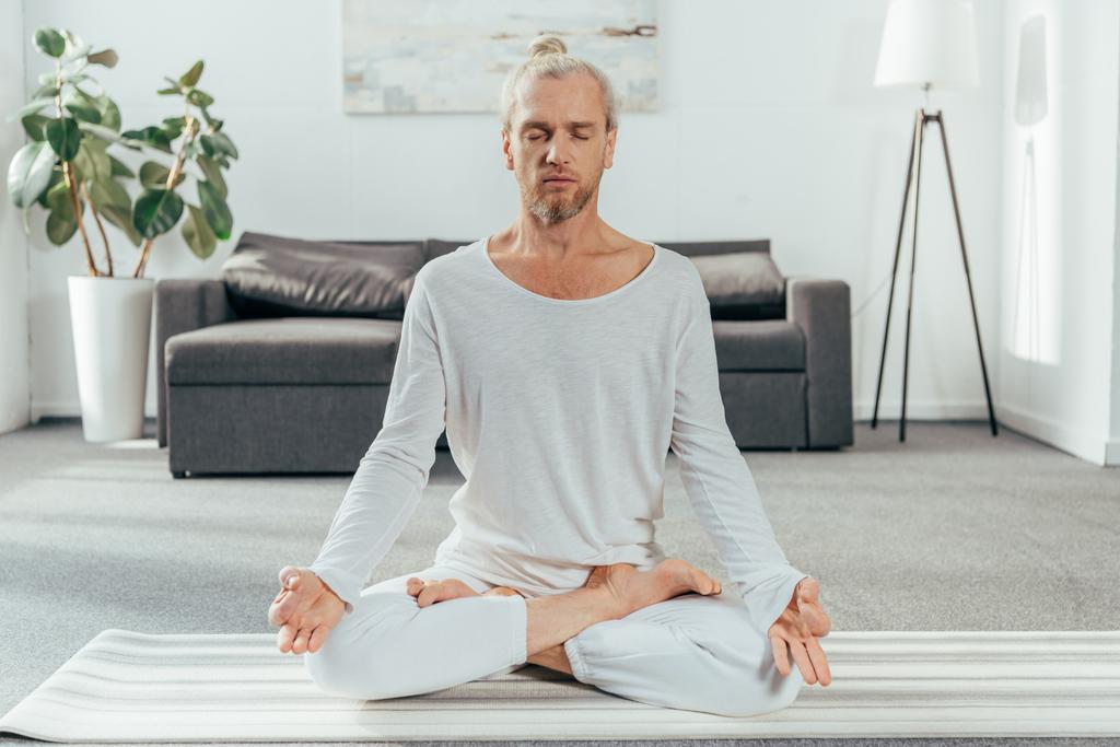 sportlicher erwachsener Mann meditiert zu Hause in Lotusposition auf Yogamatte  - Foto, Bild