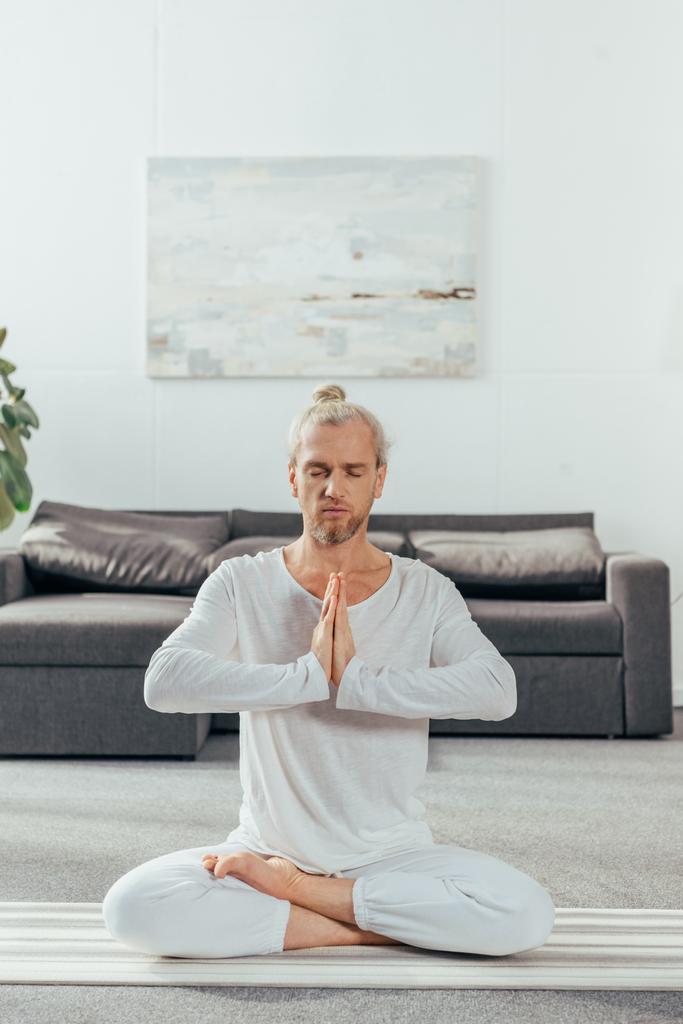 homem adulto com olhos fechados meditando em posição de lótus e gesto namaste em casa
  - Foto, Imagem