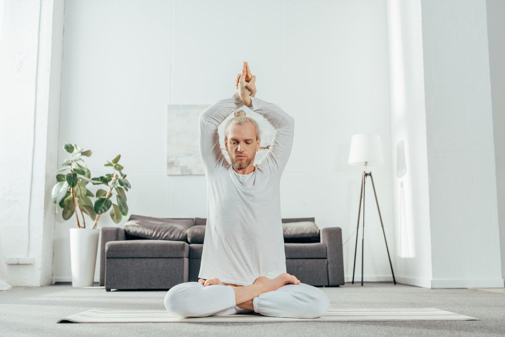 взрослый мужчина медитирует в позе лотоса со знаком мудры на коврике для йоги дома
 - Фото, изображение