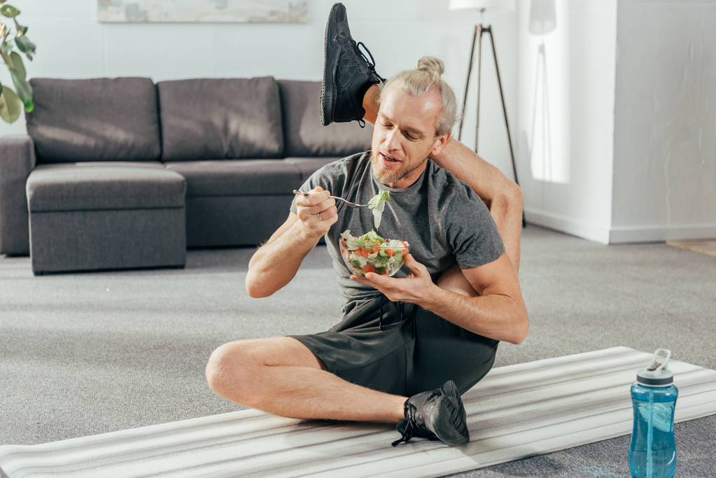 elastyczne dojrzałego mężczyzny z nogi za głowę, siedząc na matę do jogi i jedzenie warzywo Sałatka  - Zdjęcie, obraz