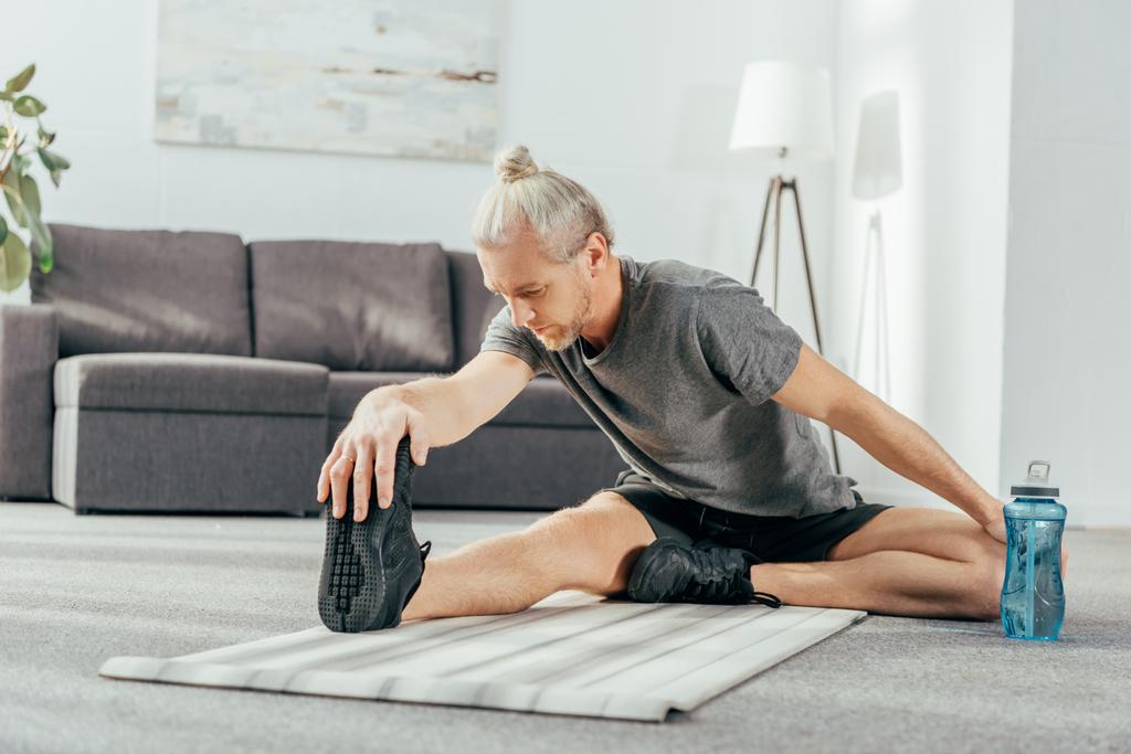 atletische man in sportkleding op yoga mat zitten en zich thuis het uitrekken - Foto, afbeelding