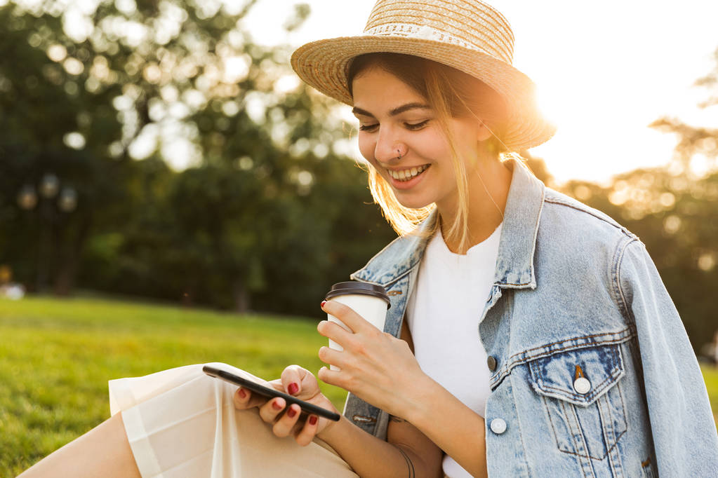 Kaunis nuori tyttö istuu nurmikolla puistossa, käyttäen matkapuhelinta, pitämällä kahvikuppia
 - Valokuva, kuva