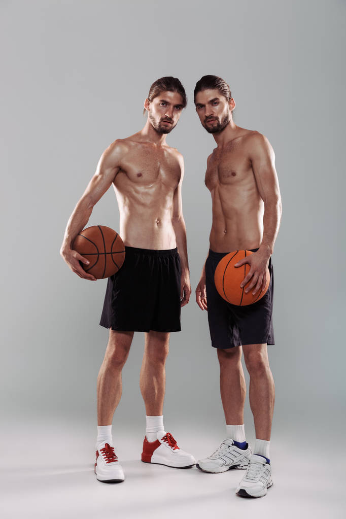 Retrato de cuerpo entero de dos hermanos gemelos musculosos sin camisa de pie aislados sobre fondo gris, sosteniendo pelotas de baloncesto
 - Foto, Imagen