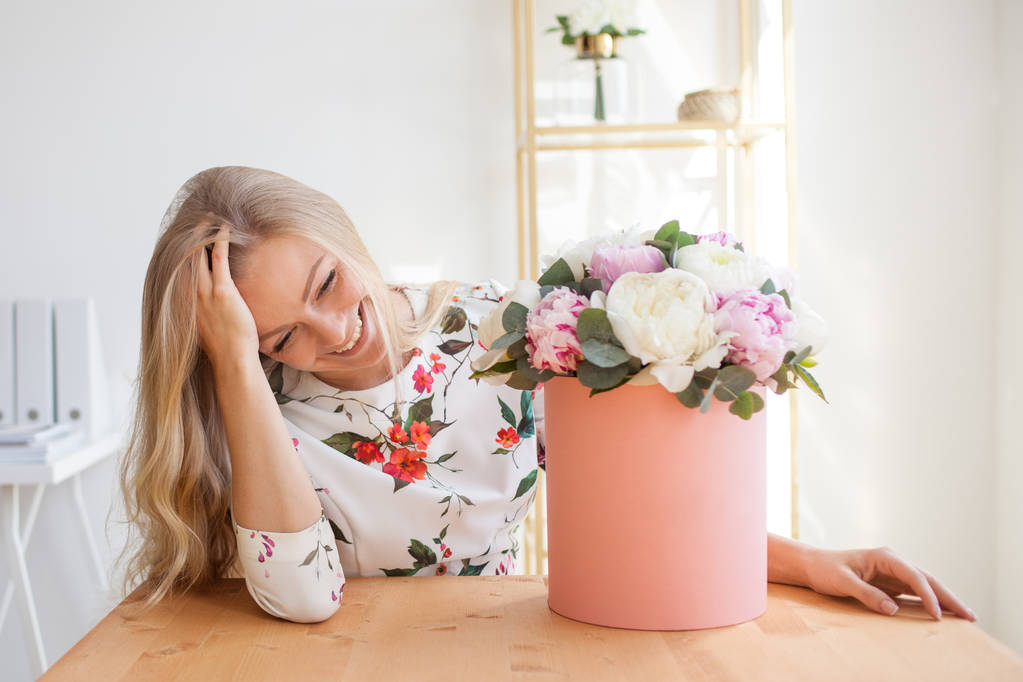 Gelukkig blonde vrouw in een modern kantoor met bloemen in een doos van de hoed. Boeket van pioenrozen. - Foto, afbeelding