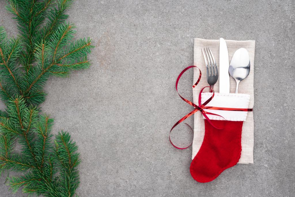 erhöhte Ansicht der Gabel, Löffel mit Messer in Weihnachtsmannsocke, umwickelt mit roter Schleife auf dem Tisch mit immergrünen Zweigen  - Foto, Bild