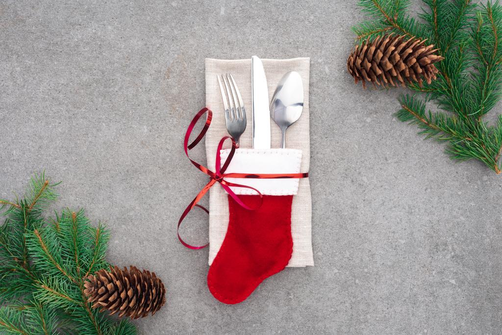 vista superior de tenedor, cuchara con cuchillo en calcetín de santa envuelto por cinta roja sobre mesa con conos de pino y ramas
  - Foto, Imagen