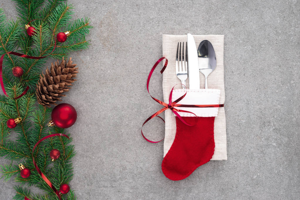 plano con tenedor, cuchara con cuchillo en calcetín de santa envuelto por cinta roja en la mesa con cono de pino y rama decorada con bolas de Navidad
  - Foto, Imagen