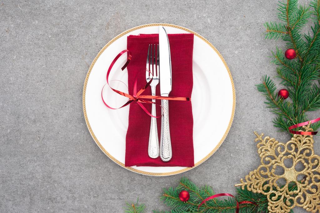 верхний вид подаваемой тарелки с вилкой и ножом, обернутый красной праздничной лентой на поверхности с вечнозелеными ветвями, украшенными рождественскими шарами и золотой звездой
  - Фото, изображение