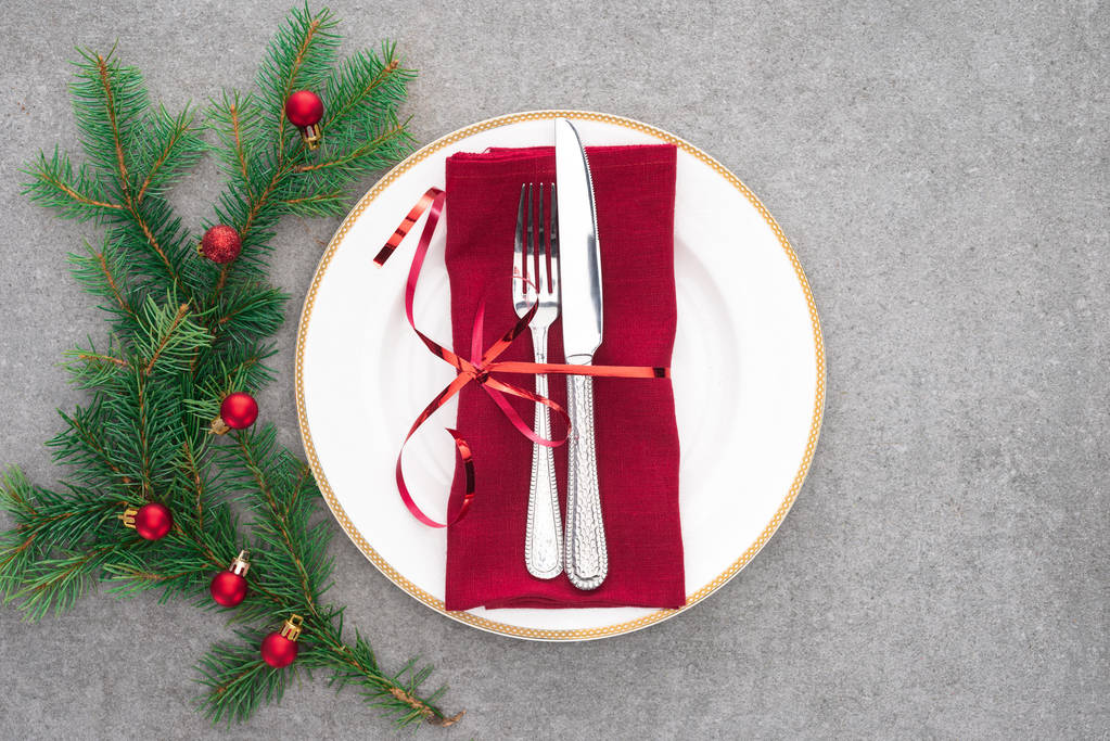 Blick von oben auf Serviertisch mit Teller, Gabel und Messer, umwickelt von festlicher Schleife am mit Weihnachtskugeln geschmückten Zweig  - Foto, Bild