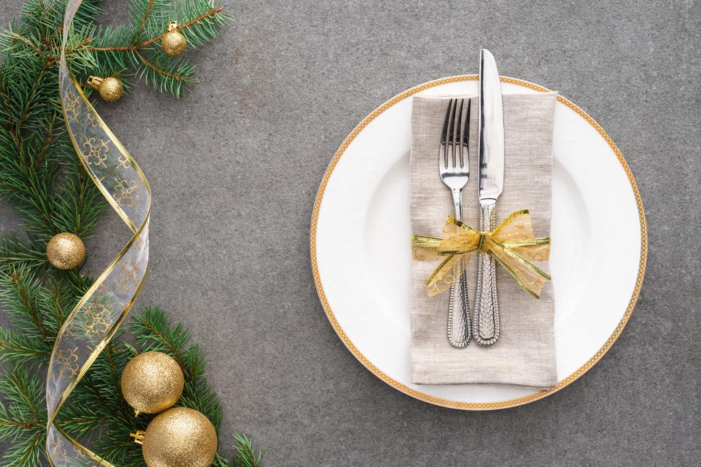 возвышенный вид обслуживаемого стола с тарелкой, вилкой и ножом, завернутым праздничной лентой рядом с веткой, украшенной золотыми рождественскими шарами
  - Фото, изображение