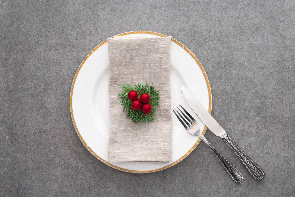 tendido plano con plato servido con tenedor, cuchillo y rama de hoja perenne decorado con bolas de Navidad en la superficie gris
 - Foto, Imagen