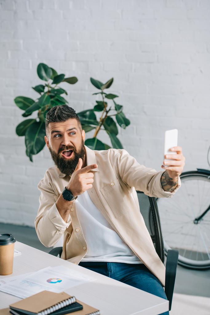 Бородатый бизнесмен делает селфи на смартфоне и жестикулирует в современном офисе
 - Фото, изображение