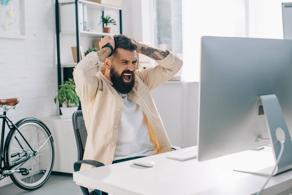 Impulsiivinen parrakas liikemies, jolla on kädet pään päällä, istuu tietokoneella ja huutaa virassa
 - Valokuva, kuva