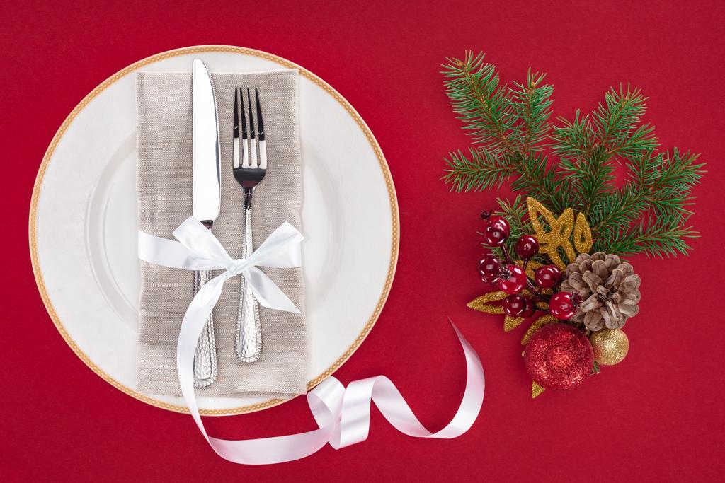 повышенный вид вилки и ножа, завернутые лентой на тарелке возле рождественских украшений, изолированных на красный
 - Фото, изображение