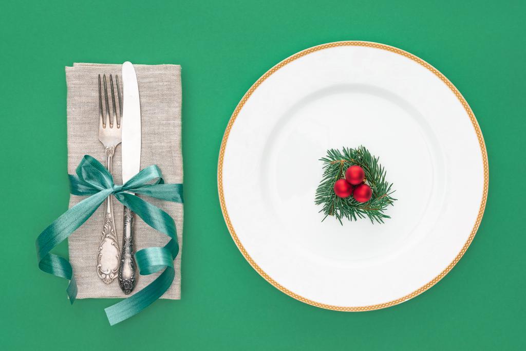 flat lay with evergreen branch and chiristmas balls on plate near fork with knife wrapped by ribbon isolated on green - Photo, Image