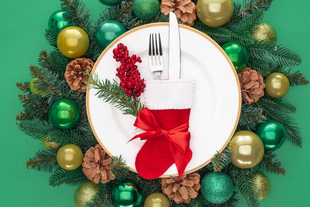 pohled shora na vidličku a nůž v santa ponožku a červené bobule na talíři uprostřed větve stálezelených stromů s vánoční koule izolované na zelené - Fotografie, Obrázek