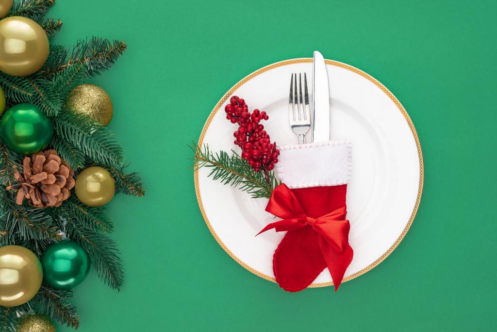 flat lay with red berries and fork with knife in christmas sock on plate near evergreen branches with christmas balls isolated on green - Photo, Image