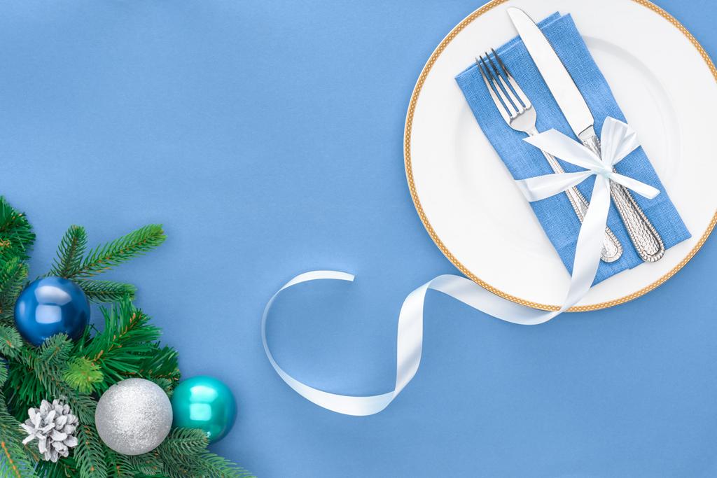 θέα από ψηλά, πιρούνι και μαχαίρι τυλιγμένο με εορταστική κορδέλα στο πιάτο κοντά αειθαλές δέντρο κλαδιά με Χριστουγεννιάτικες μπάλες που απομονώνονται σε μπλε - Φωτογραφία, εικόνα