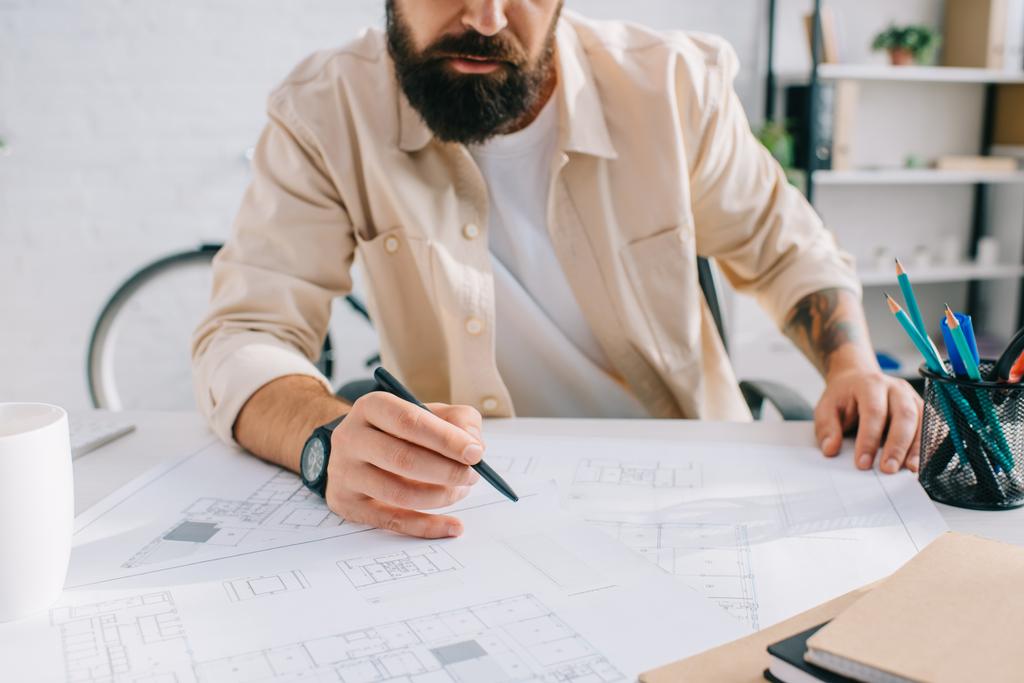 Бородатый архитектор сидит за столом и работает с чертежами
  - Фото, изображение
