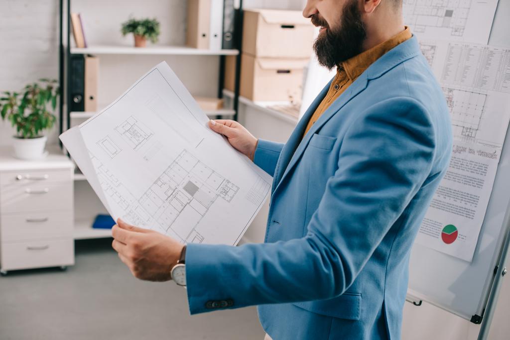 Ausgeschnittene Ansicht eines erwachsenen männlichen Architekten in blauer formaler Kleidung, der eine Blaupause hält, Flipchart verwendet und im Büro an einem Projekt arbeitet - Foto, Bild