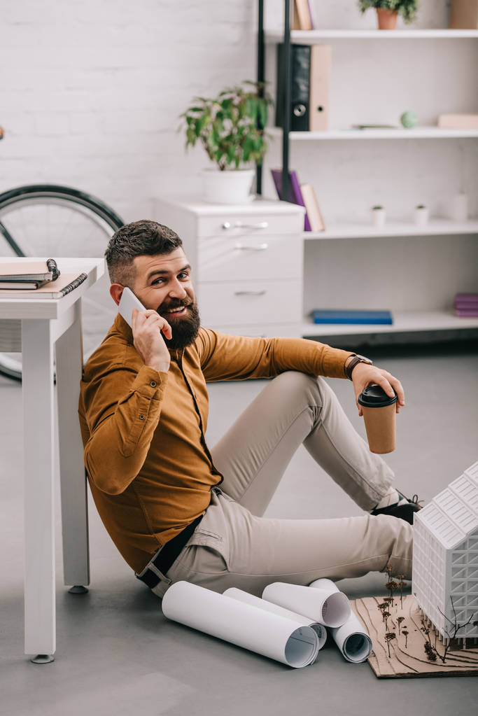 sonriente barbudo adulto arquitecto masculino hablando en smartphone, sosteniendo café para llevar y trabajando en planos en la oficina
  - Foto, Imagen