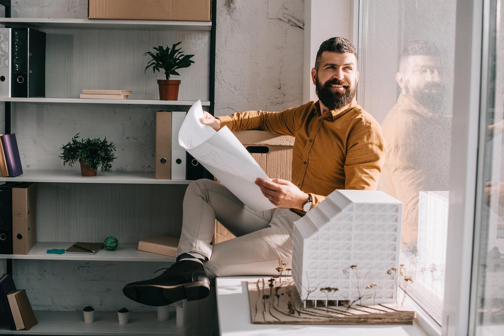 lächelnder erwachsener, bärtiger Architekt, der in der Nähe des Hausmodells sitzt, einen Entwurf in der Hand hält und im Büro an einem Projekt arbeitet  - Foto, Bild