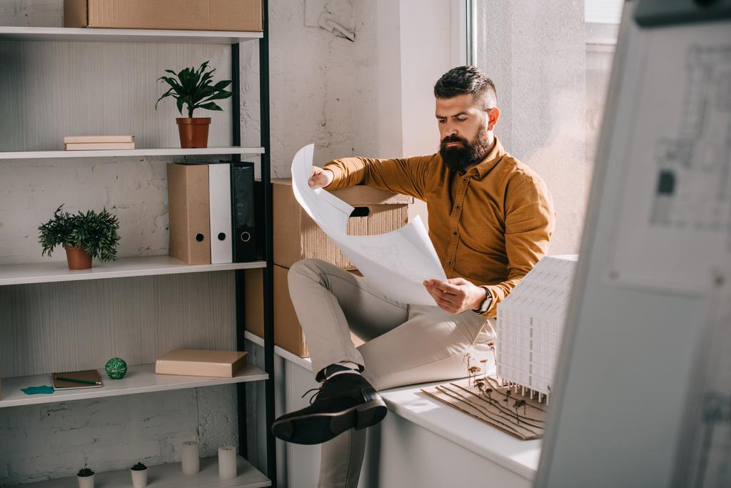 задумчивый бородатый взрослый мужчина-архитектор сидит рядом с моделью дома, держит чертеж и работает над проектом в офисе
  - Фото, изображение