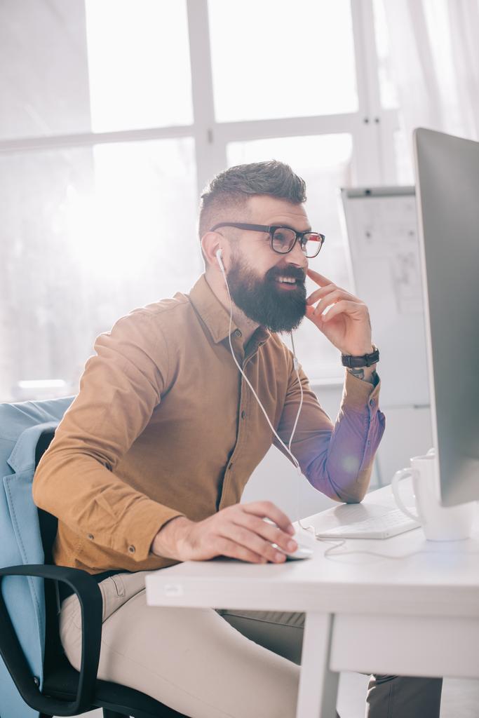 улыбающийся бородатый взрослый бизнесмен в наушниках сидит и работает за компьютерным столом в офисе
 - Фото, изображение