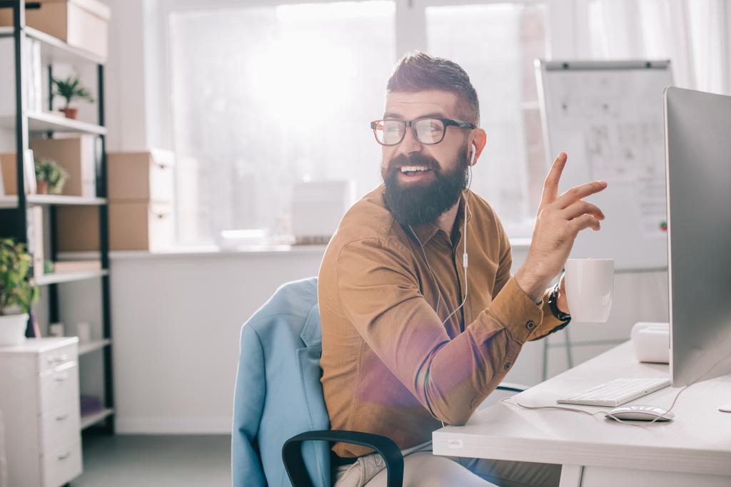 усміхнений бородатий дорослий бізнесмен в навушниках сидить і працює за комп'ютерним столом в офісі
 - Фото, зображення