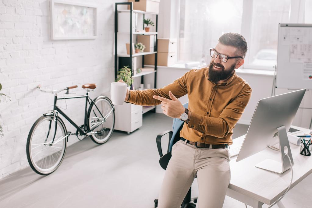 onnellinen parrakas aikuinen liikemies pitämällä kuppi kahvia ja osoittamalla sormella exitement työpaikalla
 - Valokuva, kuva