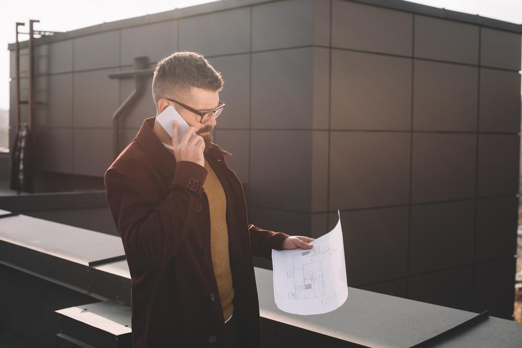 arquitecto masculino adulto serio en gafas hablando en el teléfono inteligente y sosteniendo plano en la azotea
 - Foto, Imagen