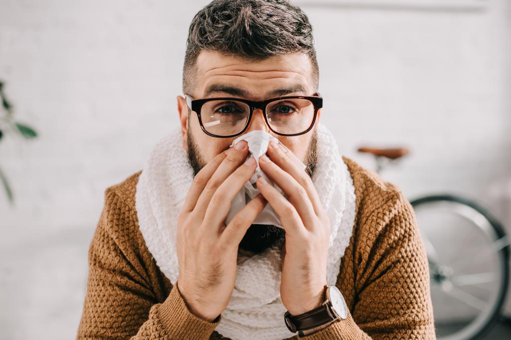 портрет больного человека в трикотажном шарфе, сидящего в офисе, чихающего и покрывающего рот тканью
  - Фото, изображение