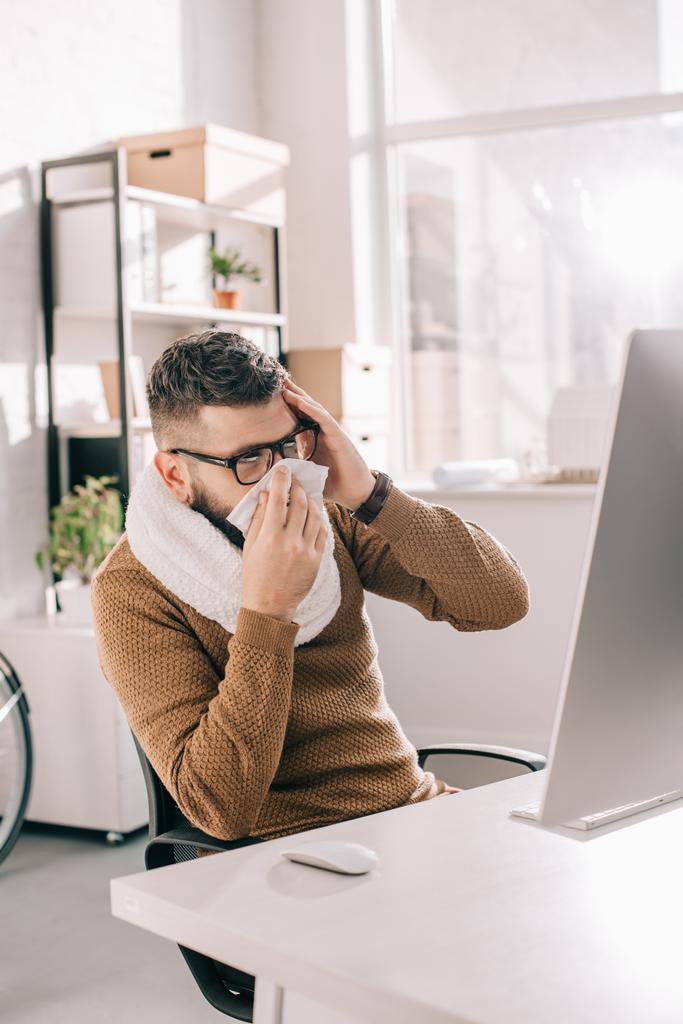 hombre de negocios enfermo en bufanda de punto sentado en la oficina, estornudando y cubriendo la boca con tejido
  - Foto, Imagen