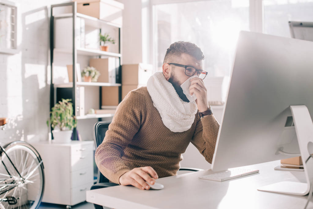 больной бизнесмен в трикотажном шарфе сидит в офисе, чихает и покрывает рот тканью
  - Фото, изображение