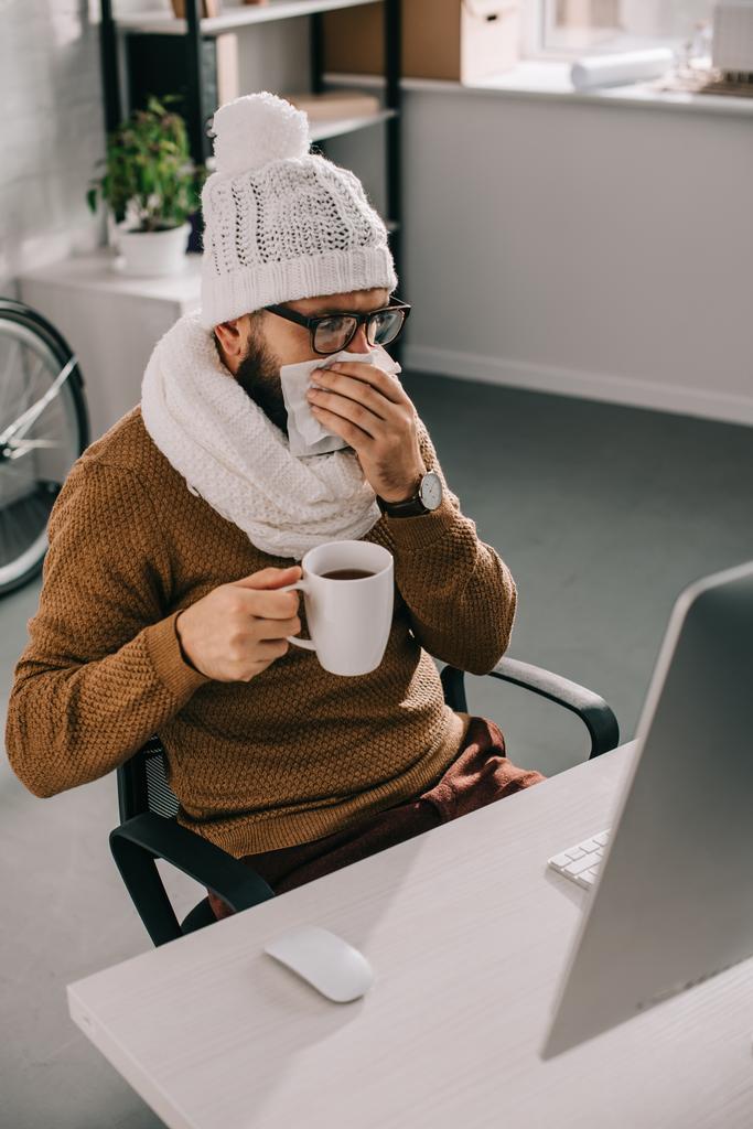 zieke zakenman in sjaal en gebreide muts zit op kantoor, bedrijf kopje thee en niezen - Foto, afbeelding