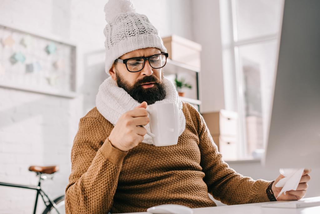 uomo d'affari malato in sciarpa e cappello a maglia che tiene tazza di tè e tessuto sul posto di lavoro
 - Foto, immagini