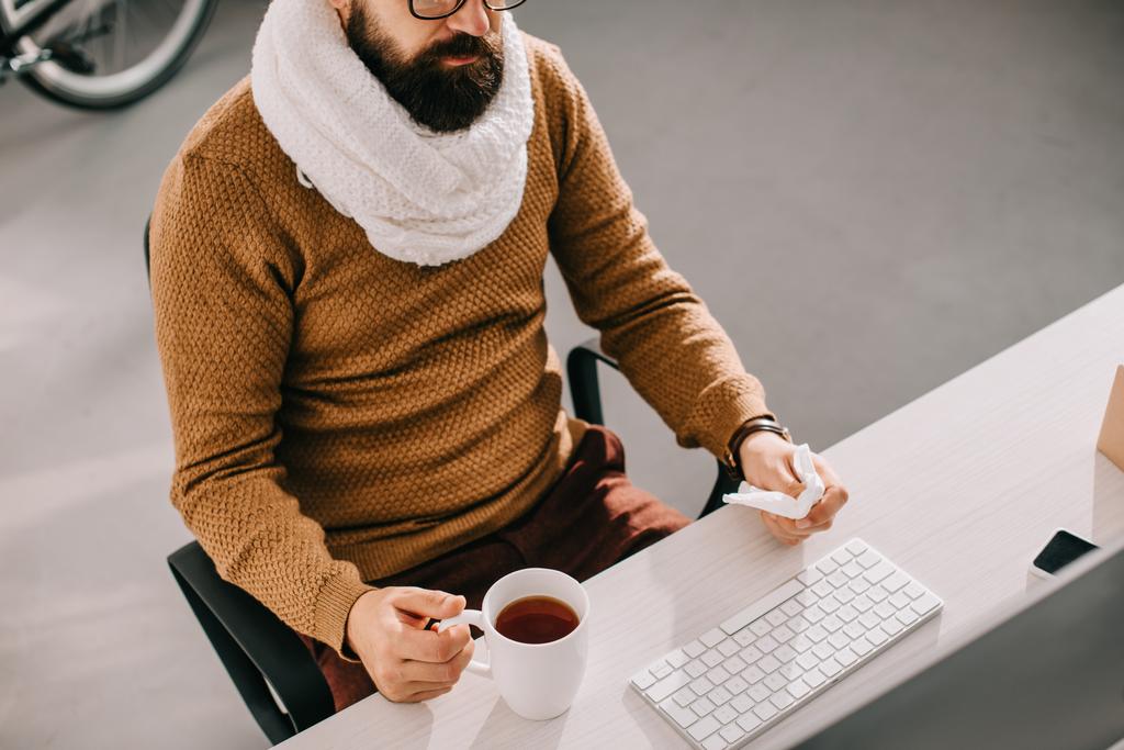 άρρωστος επιχειρηματία στο μαντήλι κρατώντας φλιτζάνι τσάι και ιστών και κάθεται στο γραφείο - Φωτογραφία, εικόνα