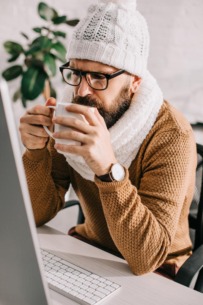 хворий бізнесмен в шарфі і в'язаний капелюх працює за комп'ютерним столом і п'є чай в офісі
 - Фото, зображення
