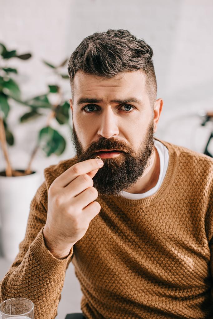 portrait of sick bearded man looking at camera and taking pill from illness  - Photo, Image