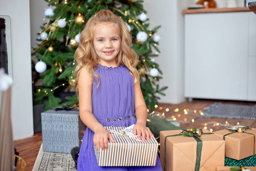 Pequena menina bonita com cabelo encaracolado loiro considera seus presentes contra o fundo da árvore de Natal. Presente de Natal, presente de Ano Novo Conceito de Natal
. - Foto, Imagem