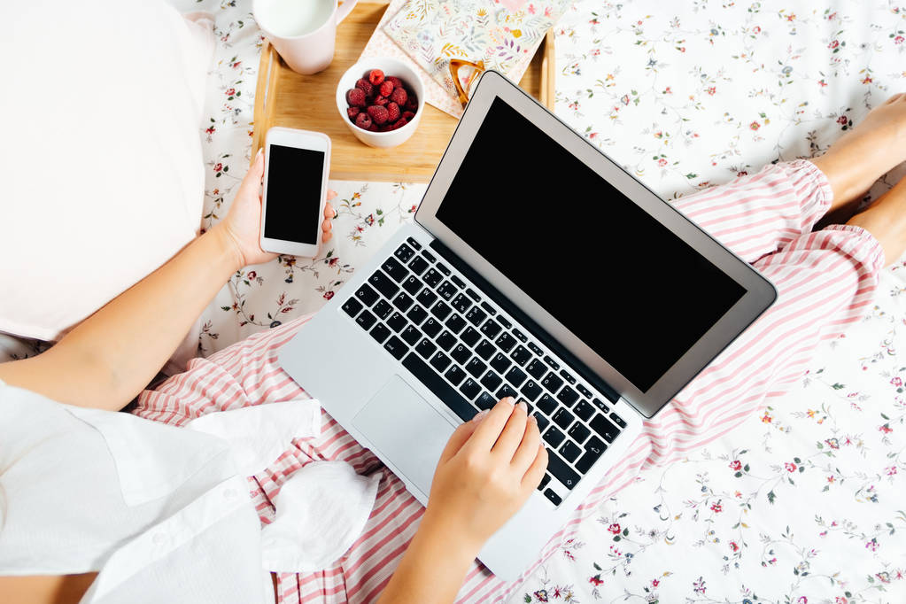 Młoda kobieta w domu w łóżku działa z laptopa i telefonu, widok z góry - Zdjęcie, obraz