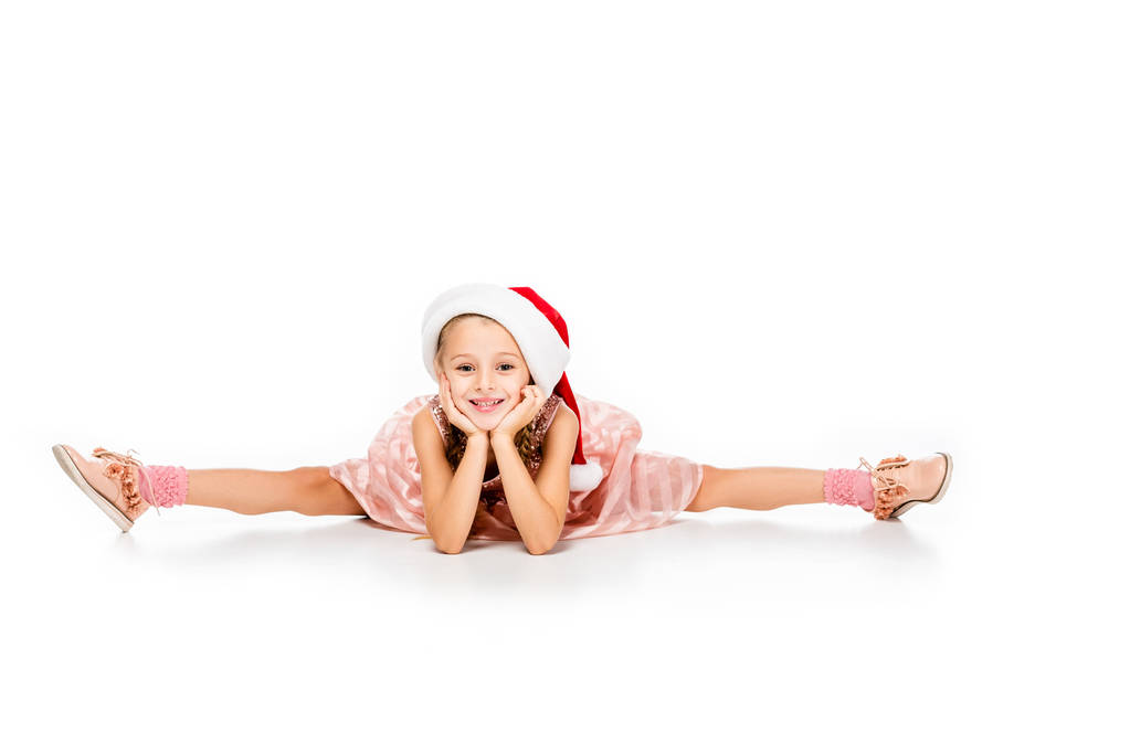 adorable little child in santa hat sitting splits on floor and looking at camera isolated on white - Photo, Image